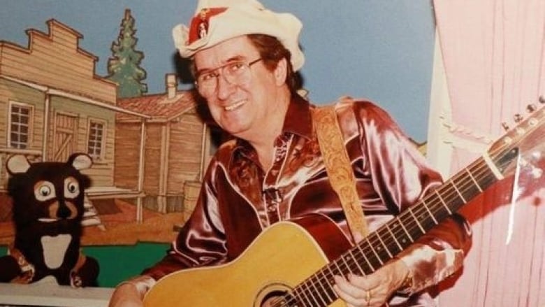 A man wearing a cowboy hat and holding a guitar stands next to a puppet bear.