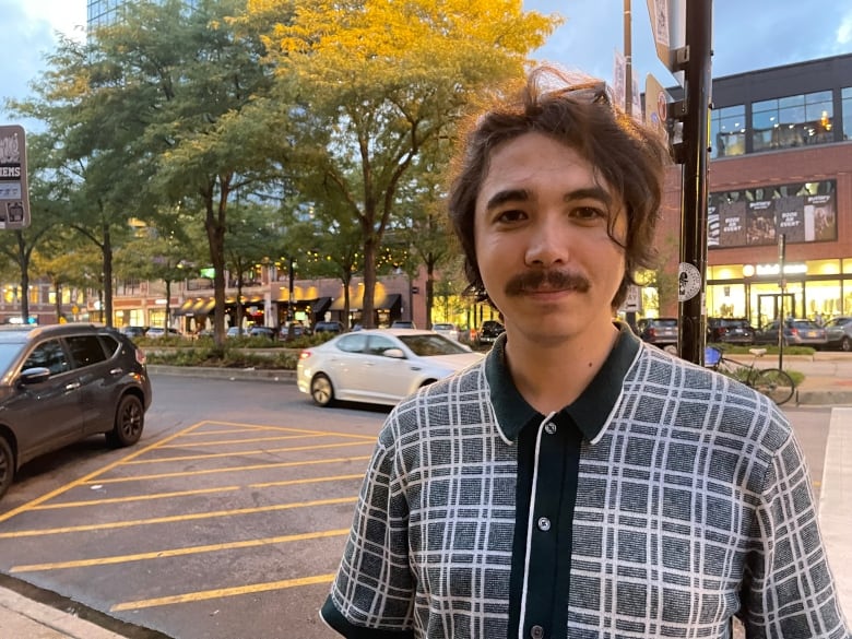 A young man with a mustache stands on a downtown street.