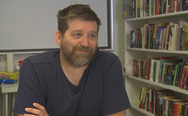 A man in a T-shirt with short blonde hair sits near a shelf of books.