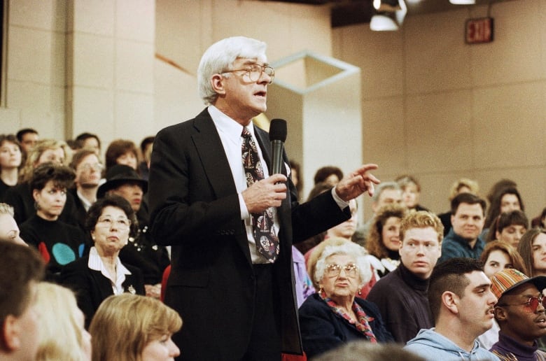 A white-haired man in glasses holds a microphone and stands among a seated audience as he speaks to someone off-camera to the right.