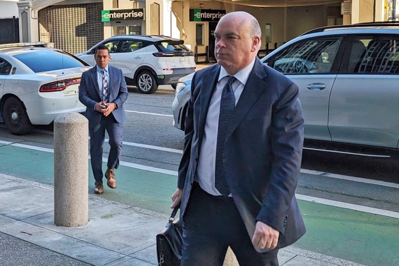 A balding man in a suit and tie carrying a black bag is shown walking on a sidewalk of a city street, seemingly toward a building.