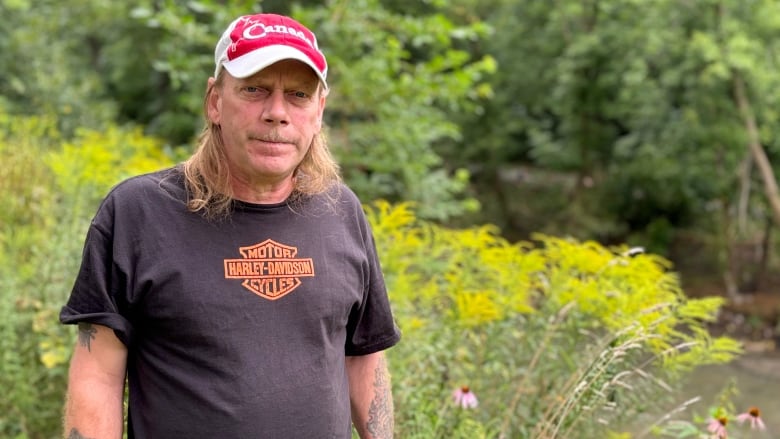 A man wearing a hat and a t-shirt stands in front of a creek.