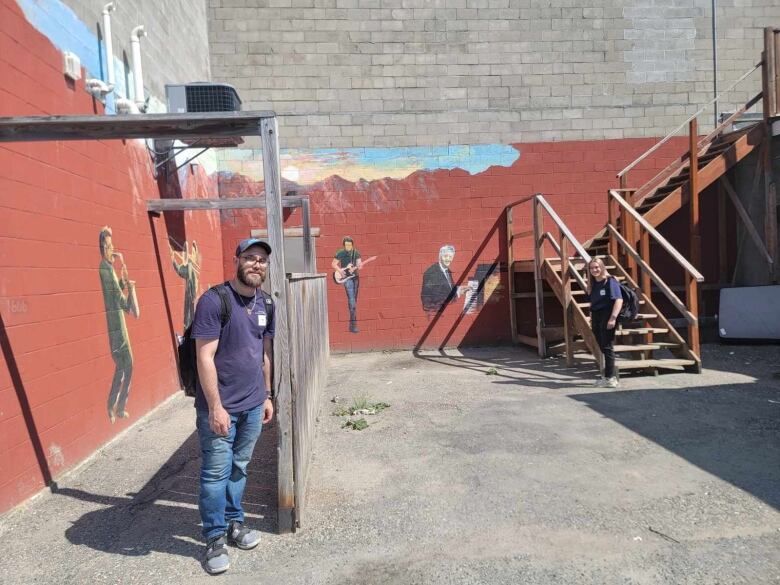 A man and woman stand in an alleyway in front of a brick wall with a mural.