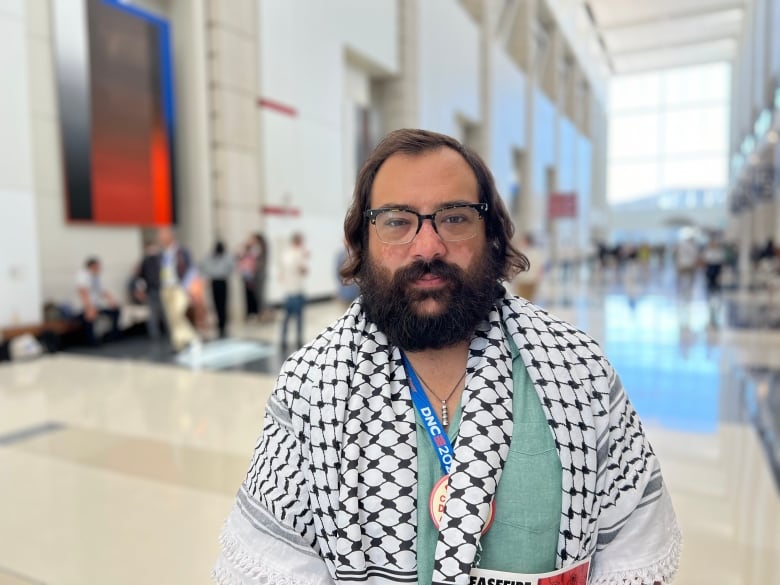 A man bearded man wears a keffiyeh scarf and a blue lanyard inside an event centre.