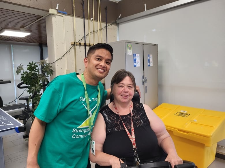 A man with a teal t-shirt smiles and stands with an arm around a woman, who is older and seated on a motorized scooter. 