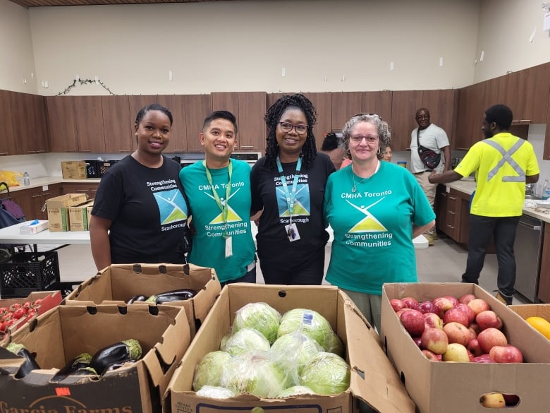 Three women and one man stand shoulder to shoulder, smiling at the camera. In front of them on tables are carboard boxes of fresh produce.