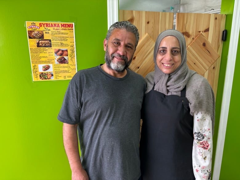 A man and woman stand in front of a green wall next to a menu for Syriana Cuisine.