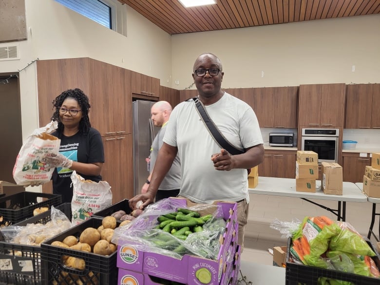 A man with a light coloured t-shirt and black glasses stands behind a table filled with fresh produce. He's looking at the camera and pointing towards the photographer.