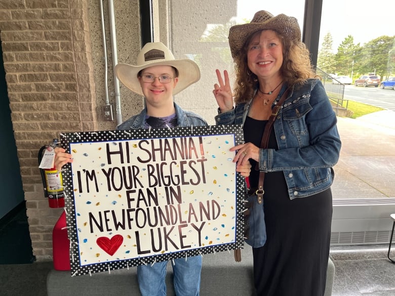 A young man holding up a sign and wearing a cowboy hat. A woman standing besides him with a cowboy hat doing the peace sign.