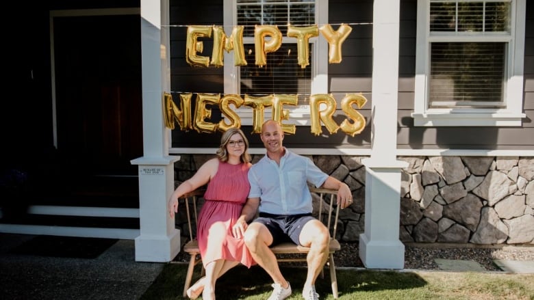 A couple in summer clothes sit on a wooden bench, with a home seen behind them and a balloon sign reading 'Empty Nesters' floating above them.