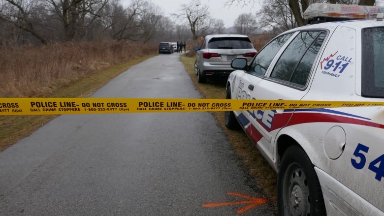 Police tape blocks off a road headed into a wooded area, with police cars lined up down the road.