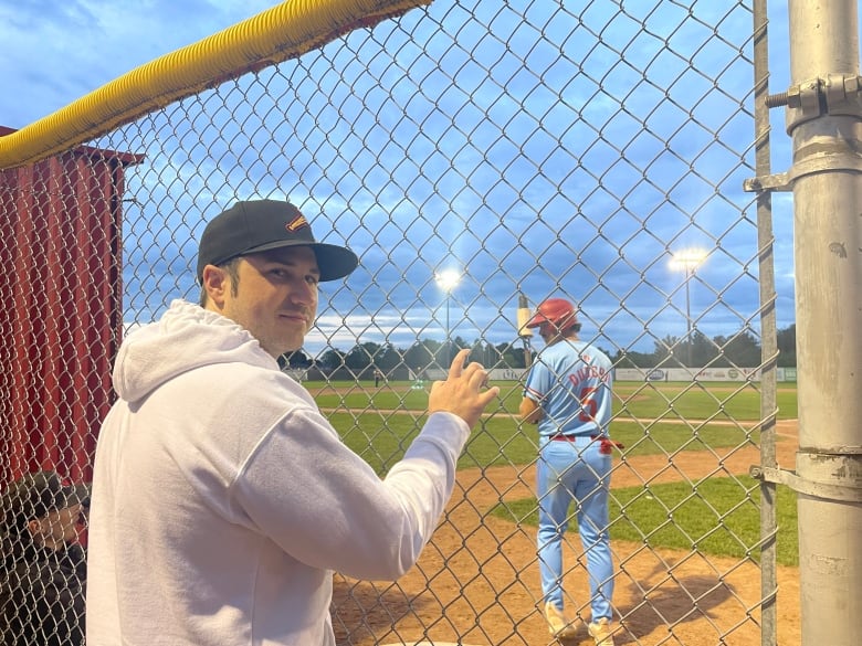 A man looking towards the camera, his hand is holding onto a metal fence, and there is a baseball player in the background.