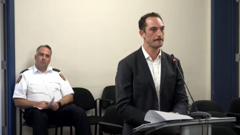 A man in a suit and open-collared white shirt stands at a podium with a man in a white police or bylaw officer uniform sitting in the spectator seats behind him.