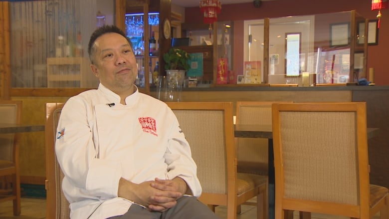 An Asian man with spiky black hair is shown sitting in a white chef's coat with his arms folded. 