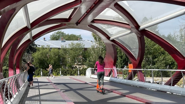 A woman wearing a bright pink shirt and grey leggings is riding an orange scooter across the Peace Bridge