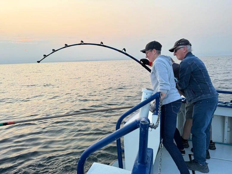 Two people stand on the edge of a boat holding onto a fishing pole which bends into the water. 