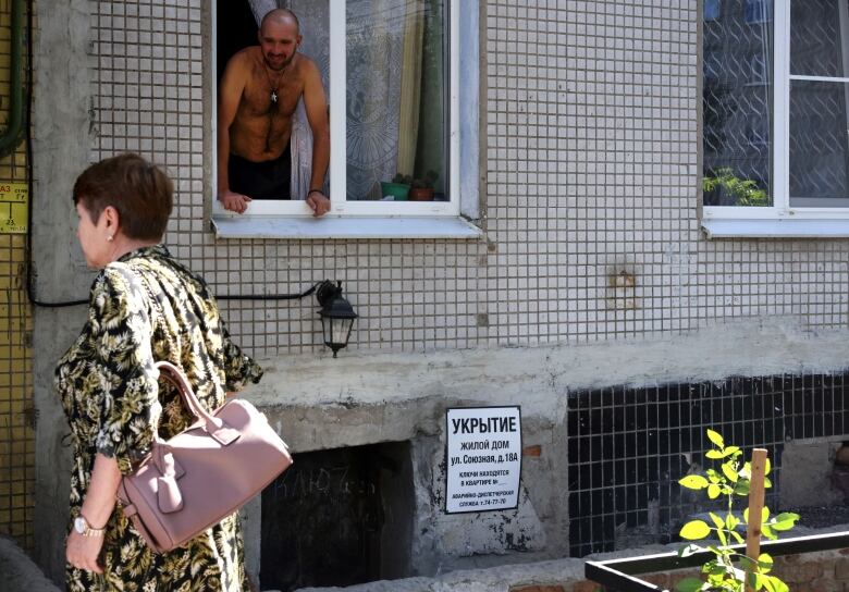 A man looks out a window in Russia's Kursk region last Friday, as a woman walks past a bomb shelter entrance.