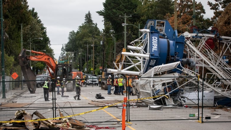 A crane lying on its side with workers standing nearby. Everything is behind a fence