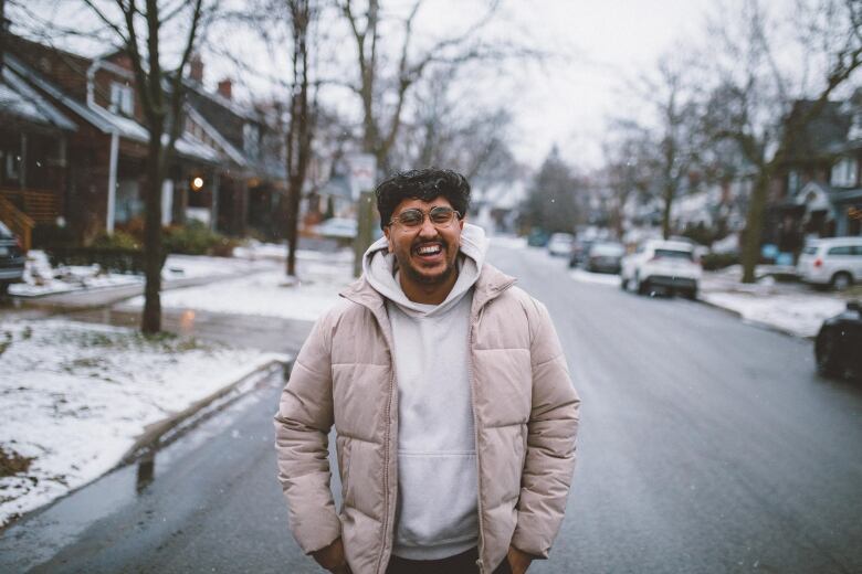 A young, laughing man wearing glasses and a winter coat stands outside on a residential street.