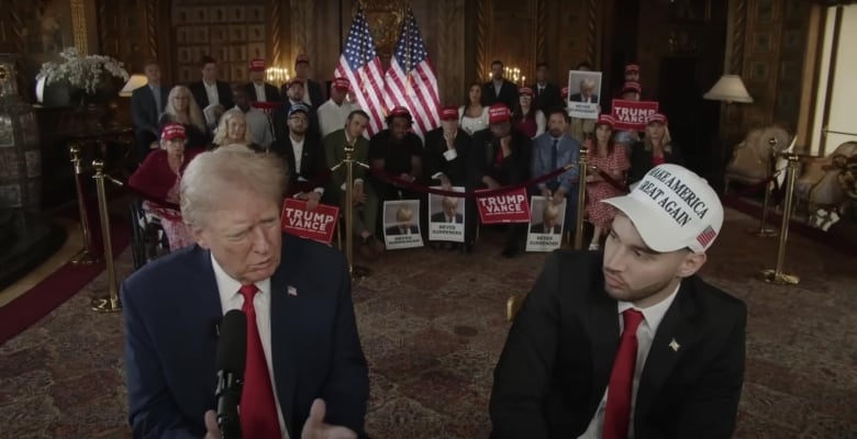 Two men sit next to each other. Behind them is a crowd of people with American flags and signs that say 'Trump Vance.'