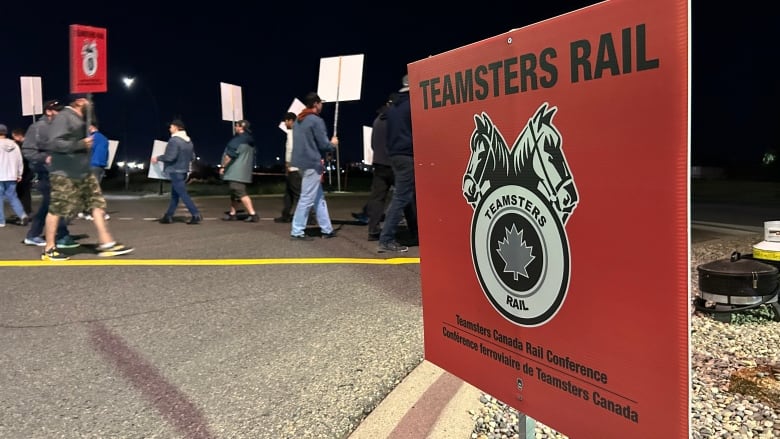 A red Teamsters sign stands before a group of people holding picket signs. 