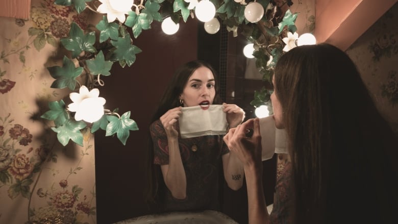 Portrait of Anna Marie Tendler blotting her lipstick in front of a flower-adorned bathroom mirror.