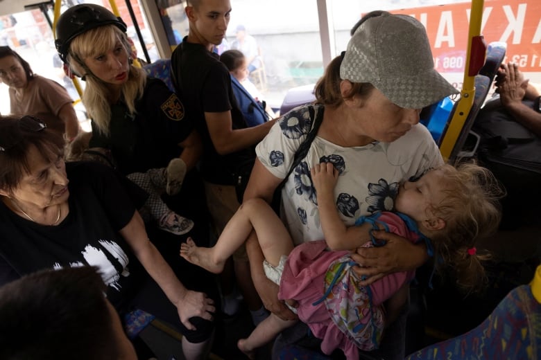 A woman in a baseball hat cradles a child who appears to be sleeping, on a bus crowded with people.