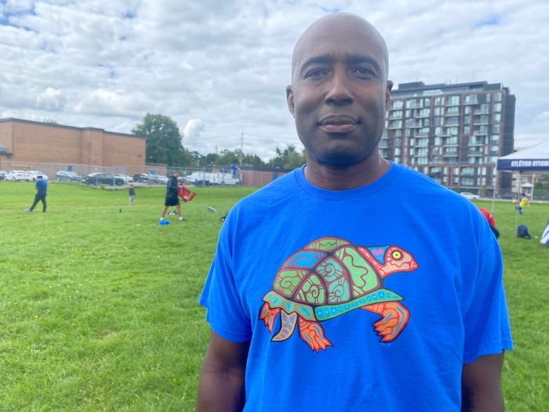 A man in a blue shirt stands on a field. Behind him, people work to set up a training session for Indigenous youths. 