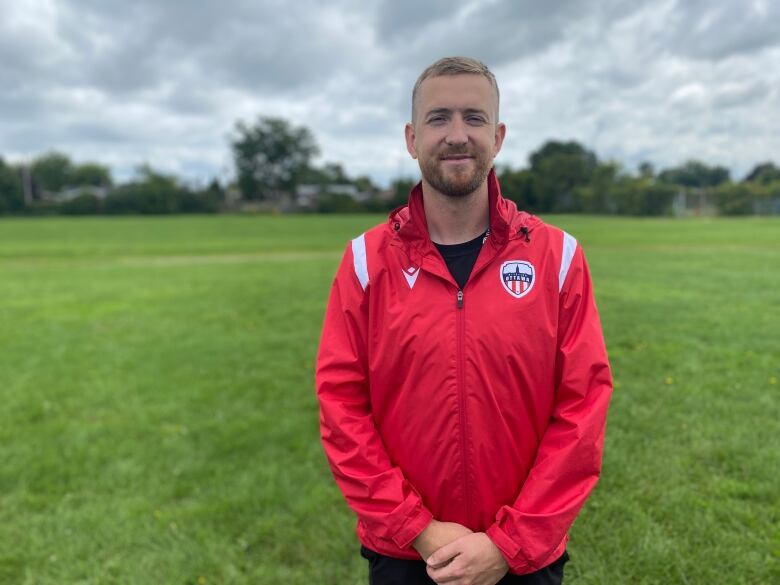 A man in a red Atltico Ottawa jacket stands with his hands crossed. 