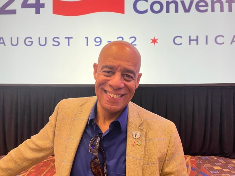 A man wearing a tan jacket and a blue dress shirt smiles.