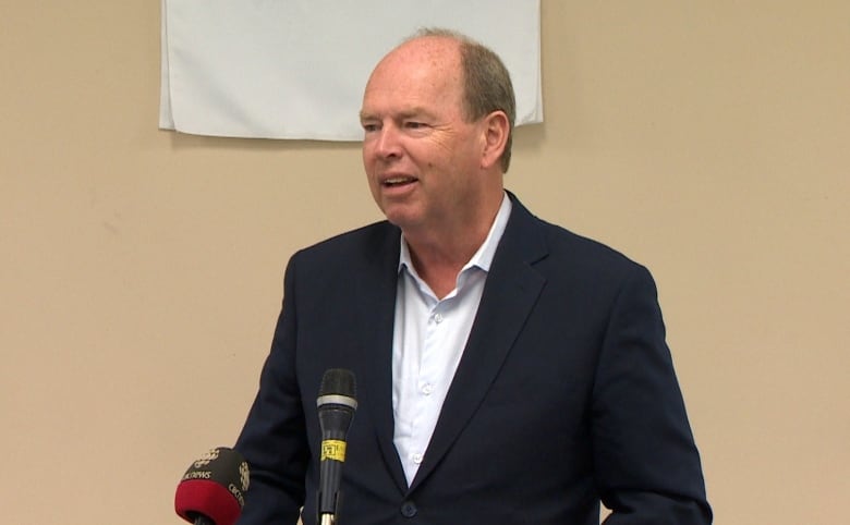 A middle aged man is seen behind a podium. Behind him is a small sign that reads 