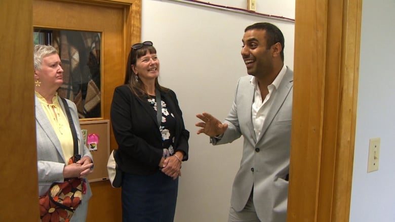 A man in a fashionable suit is seen in a hallway, addressing two women.