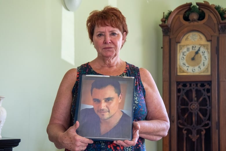 A person is seen standing in a living room, holding a photo album. On the cover of the album is a photo of a young man.