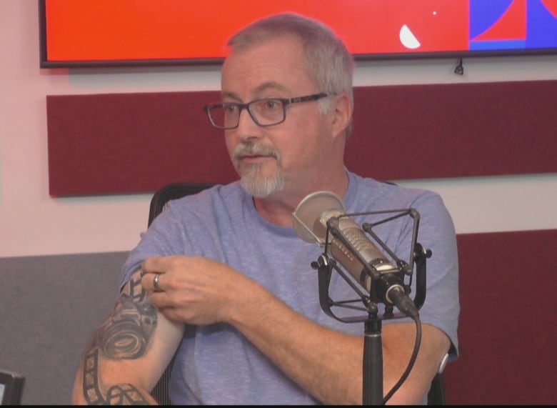 A middle-aged man lifts up the sleeve of his T-shirt to show a tattoo of a camera surrounded by a unfurling reel of film. 