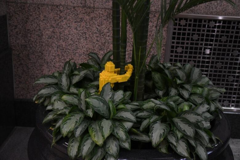 A figure made of yellow Lego bricks hugs the stalk of an indoor plant. 