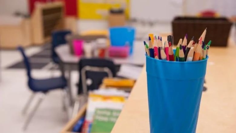 A blue cup is filled with pencil crayons. A generic classroom is blurry in the background. 