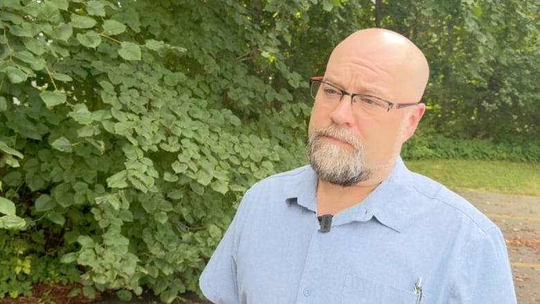 A man with a blue button-up shirt and glasses stands outside in front of greenery. 