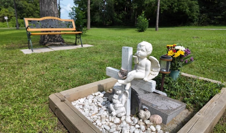 A memorial where an angel sits on a cross in a small garden with a small headstone. A bench can be seen in the background.