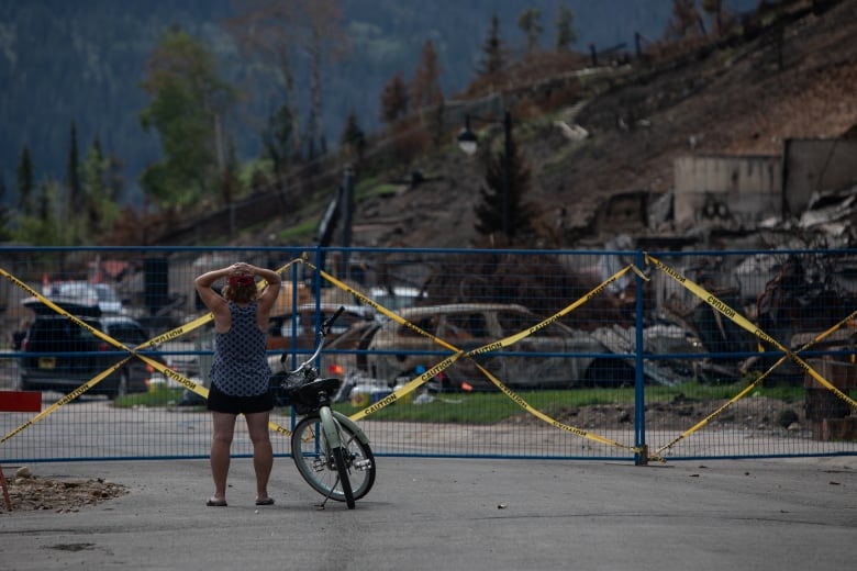 A woman with a bike stands before burned rubble surrounded by blue safety fencing. 