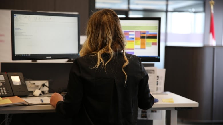 A blonde woman sits at a desk with her back to the camera.