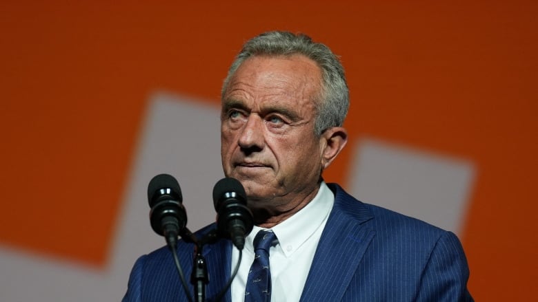 A grey-haired man in a navy suit and blue tie stands at a podium. The background behind him is red.