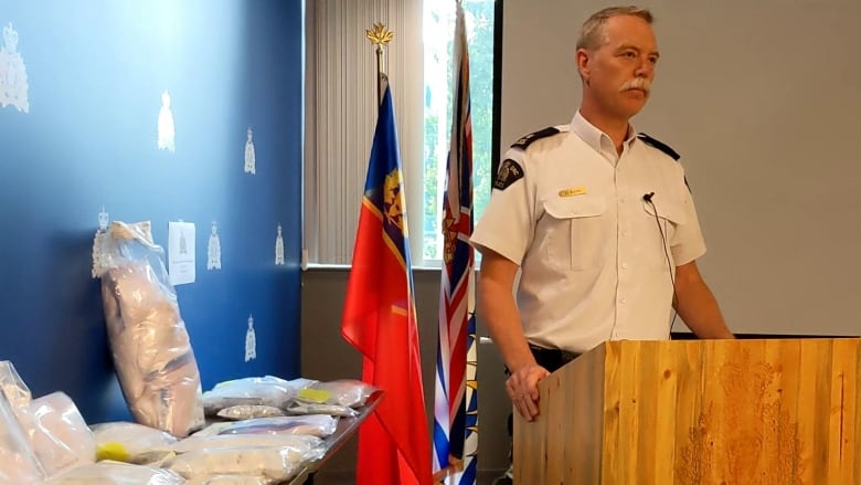 A man stands at a podium in an RCMP detachment with a large number of bagged drugs beside him.