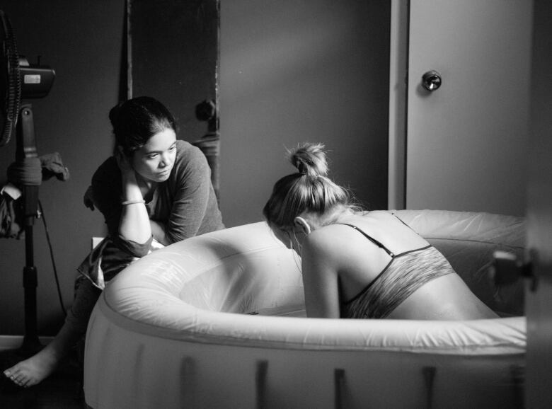 A woman labours in an inflatable birthing pool while another woman sits beside the pool.