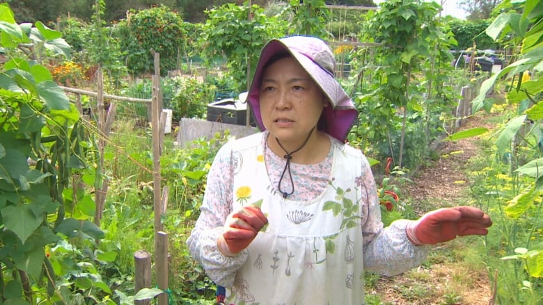 A woman dressed in her garden clothes with gloves and a hat poses in front of her garden. 