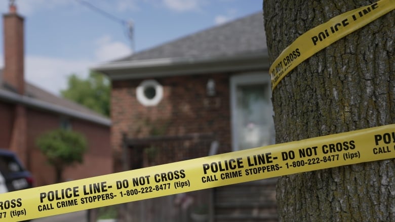 Police tape is wrapped around a tree in the foreground, with a brick house in the background. The house is blurred. It's a sunny day