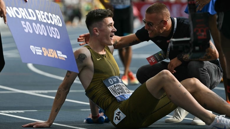 A runner smiles while sitting on the track, while a large sign that says 'WORLD RECORD 50 000 USD' is displayed behind him.