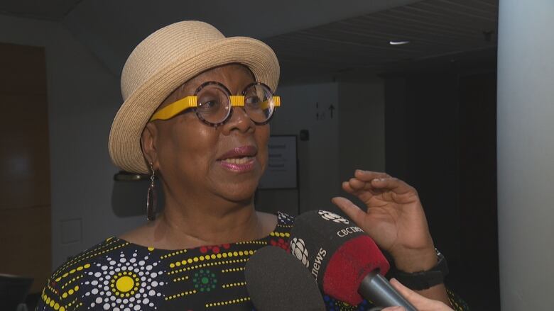 A middle aged woman in a hat and rimmed glasses speaks into two reporters' microphones inside a theatre lobby. She is visible from the shoulders up