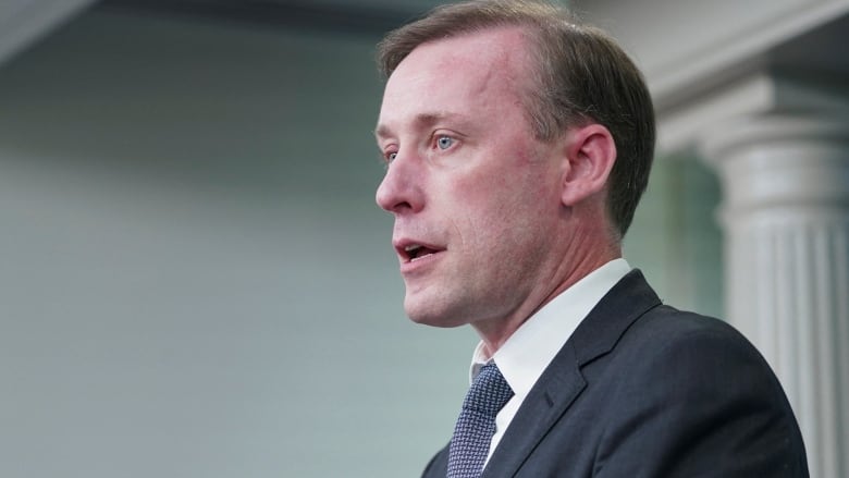 A man with short brown hair, wearing a blue jacket and tie, speaks to an audience.