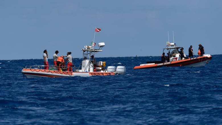 Two orange and white rescue boats with scuba divers and other people standing on them are floating in the ocean. 