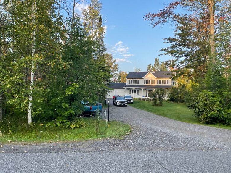 A two-storey single family home with trees on three sides and police cars on the gravel driveway.